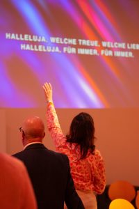 Ansicht aus dem Publikum: Eine Frau im bunt gemusterten Oberteil streckt ihren rechten Arm nach oben in Richtung Bühne. Neben ihr steht Hermann Schneider. Im Hintergrund ist auf einer Leinwand der Liedtext "HALLELUJA, WELCHE TREUE, WELCHE LIEB" und "HALLELUJA, FÜR IMMER, FÜR IMMER." zu lesen. Die Leinwand ist farbig beleuchtet.