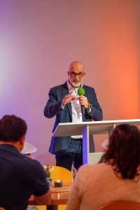 Hermann Schneider spricht an einem weißen Stehpult in ein grünes Mikrofon. Er trägt ein dunkles Sakko und ein weißes Hemd. Im unscharfen Vordergrund sind die Rücken und Hinterköpfe von zwei Zuhörern zu sehen. Der Hintergrund wird von pinkem und orangefarbenem Licht angestrahlt.