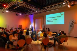 Weitwinkelaufnahme eines Veranstaltungssaals. Das Publikum sitzt an runden Tischen mit orangefarbenen Stühlen und schaut zur Bühne, wo Hermann Schneider an einem Stehpult spricht. Auf einer großen Leinwand wird der Text "Apostolic Encounter Starter" und "Apostolic Encounter Commercials" projiziert. Der Raum hat eine sichtbare Deckenkonstruktion und ist stimmungsvoll in bunten Farben beleuchtet. Rechts im Bild steht ein Beachflag-Banner.