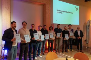 Ein Gruppenfoto von zehn Männern auf einer Bühne. Neun von ihnen halten hellblaue Zertifikate mit dem Logo "Apostolic Encounter" in die Kamera. Das Gesicht eines Mannes ist verpixelt. Ganz rechts steht Hermann Schneider mit einem Mikrofon in der Hand. Auf einer Leinwand im Hintergrund steht der Text "Sendung Apostolic Encounter 2025/26".